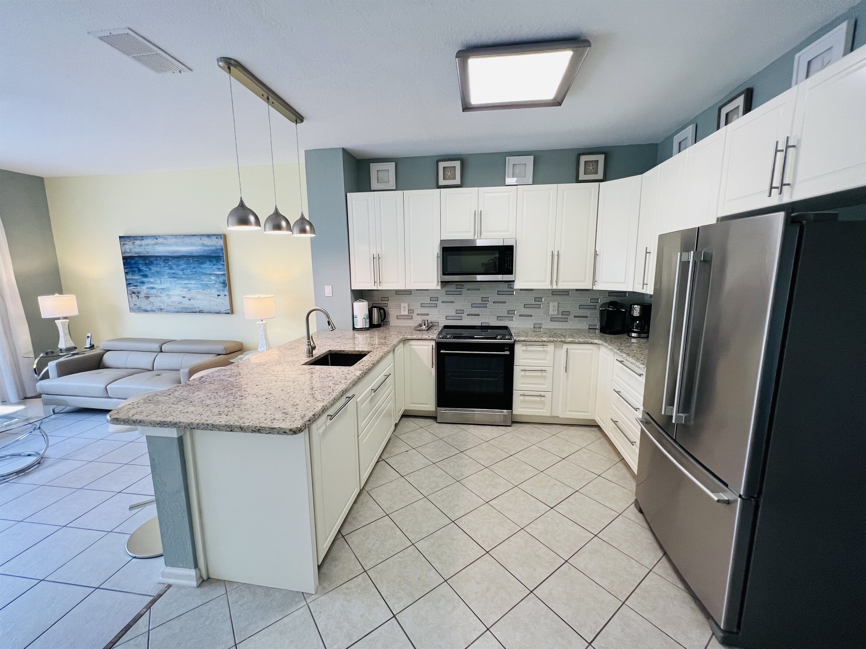 A modern kitchen with white cabinets, a stainless steel refrigerator, a microwave, and a stove. There is a dining area with a table and chairs.