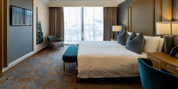 A hotel room with a large bed, a desk with a lamp, a chair, a small table, and a window with curtains. - Southern Sun Silverstar (Krugersdorp)