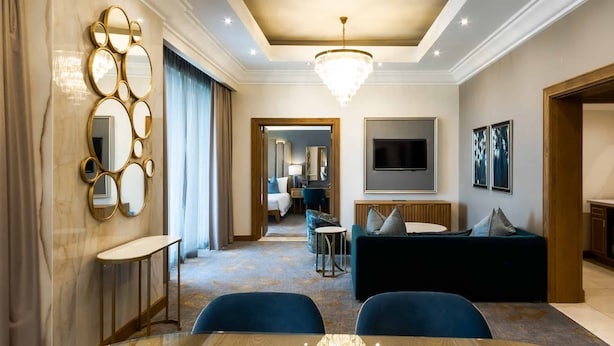 Una habitación de hotel moderna con comedor, sala de estar con sofá y televisor, y un pasillo que conduce a un baño.