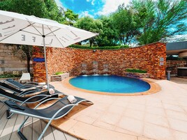 Piscina com espreguiçadeiras e um guarda-sol, cercada por paredes de pedra e vegetação.