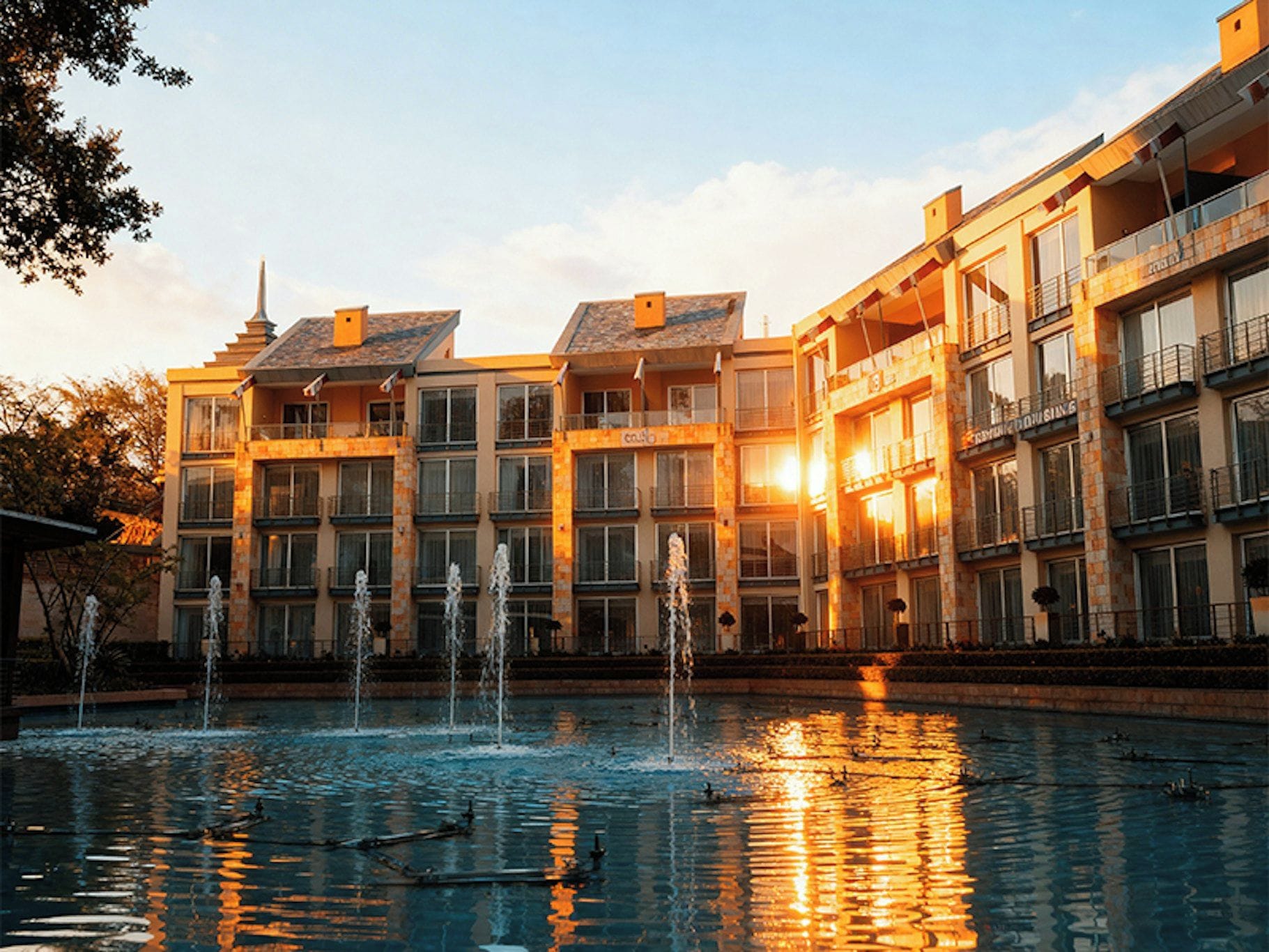 A hotel with multiple balconies, a swimming pool with fountains, and a sunset in the background.
