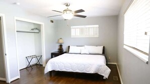 Un dormitorio con cama, ventilador de techo, mesita de noche, lámpara, ventana con persianas y un vestidor.