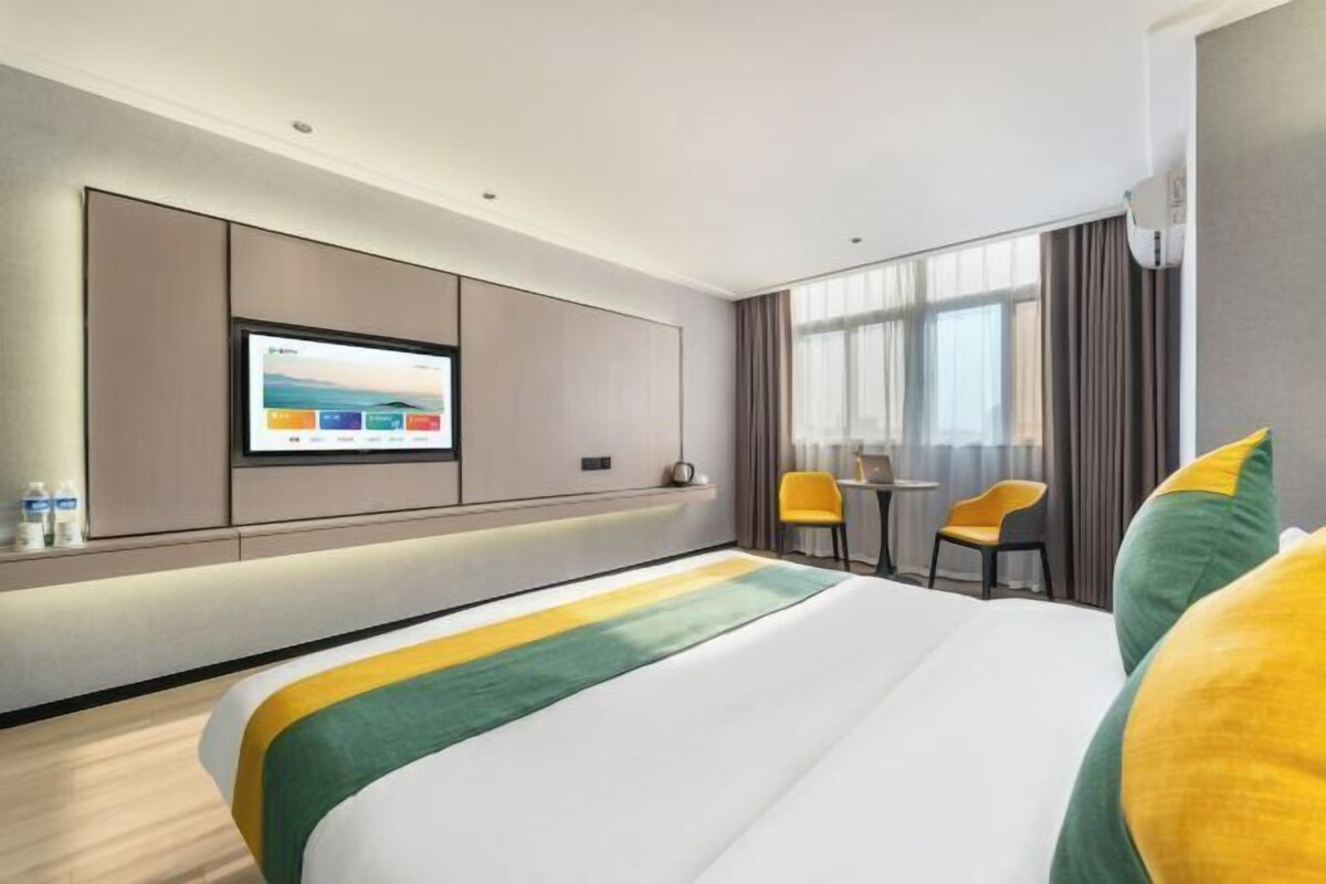 Ein modernes Hotelzimmer mit einem großen Bett, einem flachbildfernseher an der Wand, einem kleinen Tisch mit zwei Stühlen und einem großen Fenster mit Vorhängen.