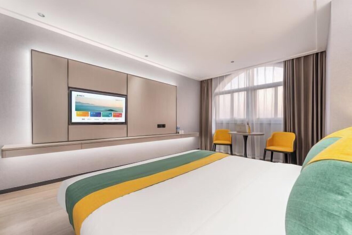 Ein Hotelzimmer mit einem Bett, einem an der Wand befestigten Fernseher, zwei Stühlen und einem mit Vorhängen versehenen Fenster.