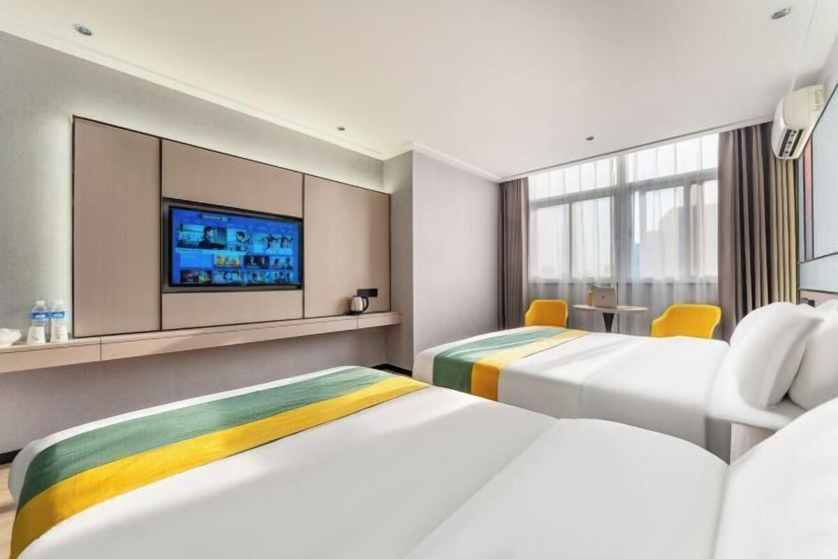 Ein Hotelzimmer mit zwei Betten, einem an der Wand montierten Fernseher, einem Fenster mit Vorhängen und einem kleinen Tisch mit Stühlen.