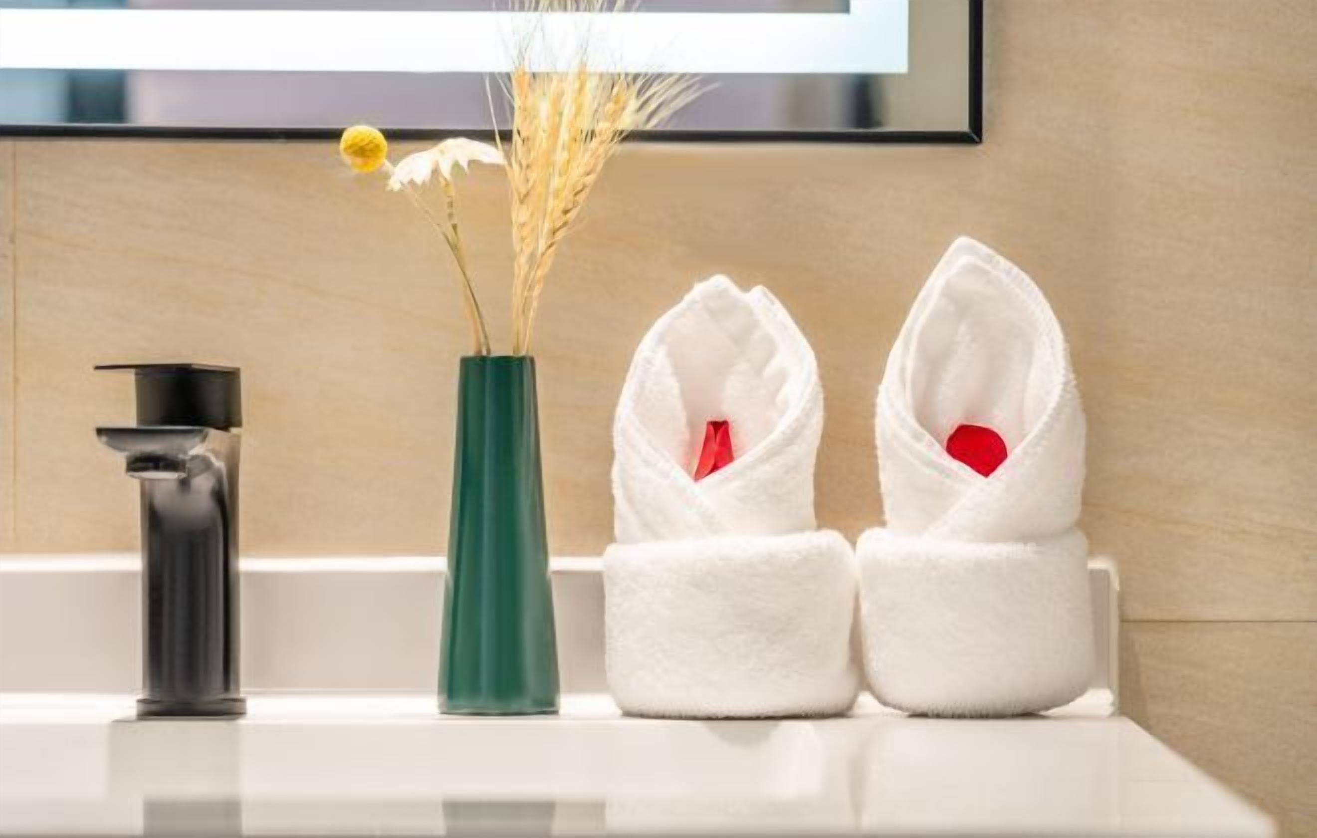 A bathroom with a sink, a green vase with dried flowers, and two white towels with red heart-shaped tags.