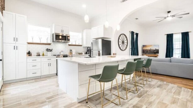 A modern kitchen with white cabinets, a central island with bar stools, and a dining area with a grey sofa and blue curtains.