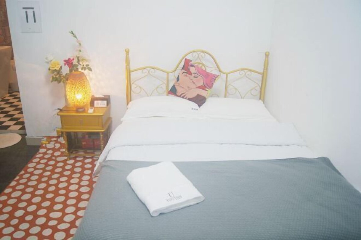 A neatly made bed with a white pillow and a folded white towel on a grey bedspread. A decorative lamp with flowers is on a small yellow table beside the bed.