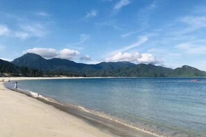 A beach with clear water, a sandy shore, and mountains in the background.