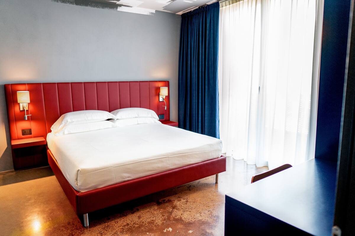 A hotel room with a red bed, white linens, a nightstand with a lamp, and a large window with blue curtains.