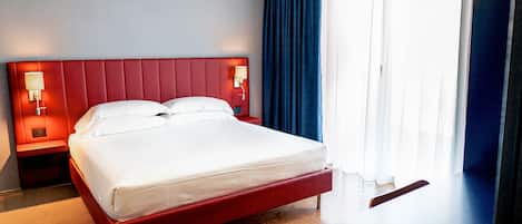 A hotel room with a red bed, white linens, a nightstand with a lamp, and a large window with blue curtains.