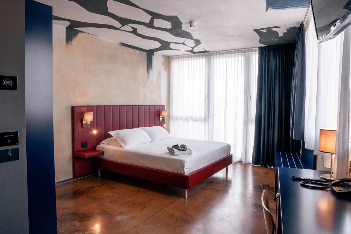 A hotel room with a bed, bedside table, lamp, and a view of the outside through a window.