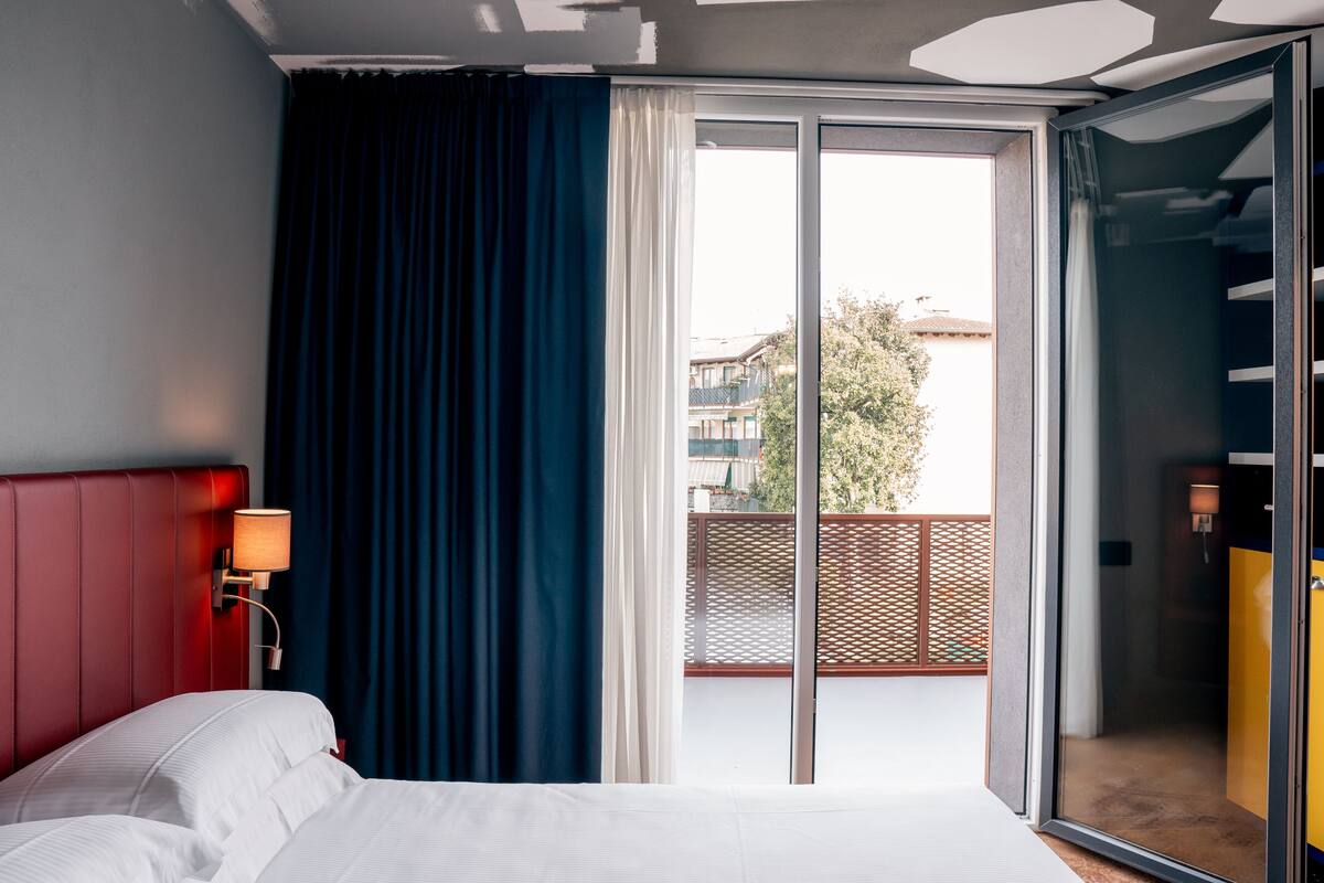 A hotel room with a bed, a nightstand, a lamp, a sliding glass door to a balcony, and a view of the outside.