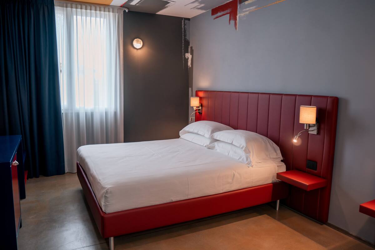 A hotel room with a red bed, a bedside table with a lamp, a window with curtains, and a wall-mounted light.