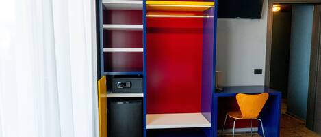 Una habitación con una estantería azul y roja, un escritorio pequeño con una silla naranja y un microondas.