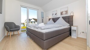 Un dormitorio moderno con una cama grande, un sillón gris y vistas a edificios a través de un amplio ventanal.