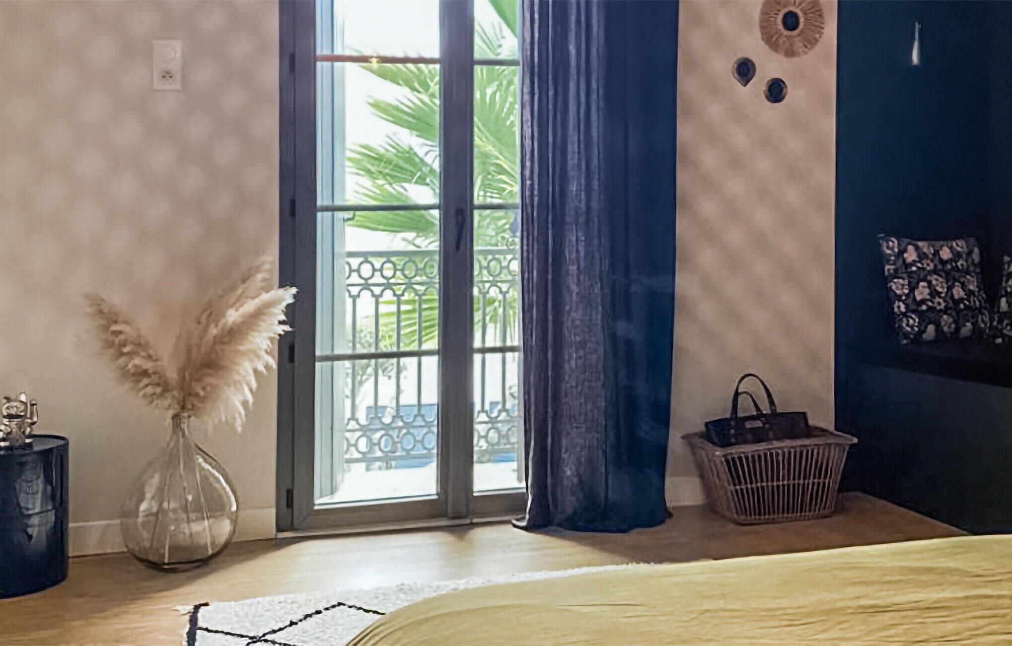 Een kamer met een grote schuifglazen deur naar een balkon, een bed met een wit hoofdeinde, een kleine vaas met pluisgras, een rieten mand en een wand met een ronde decoratie.