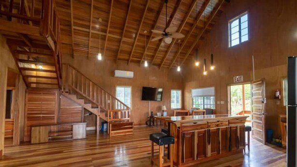 Une pièce spacieuse avec un plancher en bois, un îlot central doté de tabourets de bar, un escalier et un ventilateur de plafond.