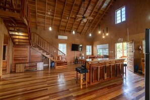 Un'ampia stanza con pavimento in legno, un'isola centrale con sgabelli da bar, una scala e una ventola a soffitto.