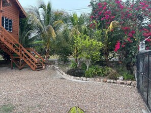 A wooden building with a staircase, surrounded by gravel, palm trees, and flowering plants.