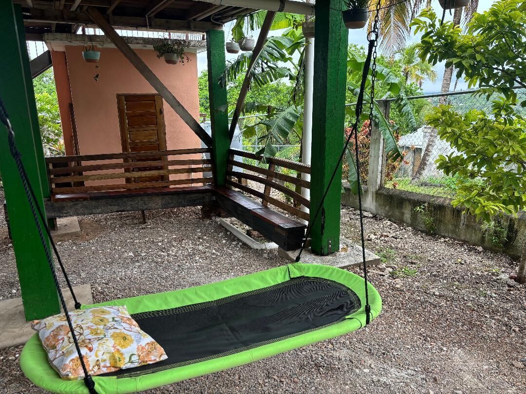 Una hamaca verde con cojín, un banco de madera y un edificio con una puerta al fondo.