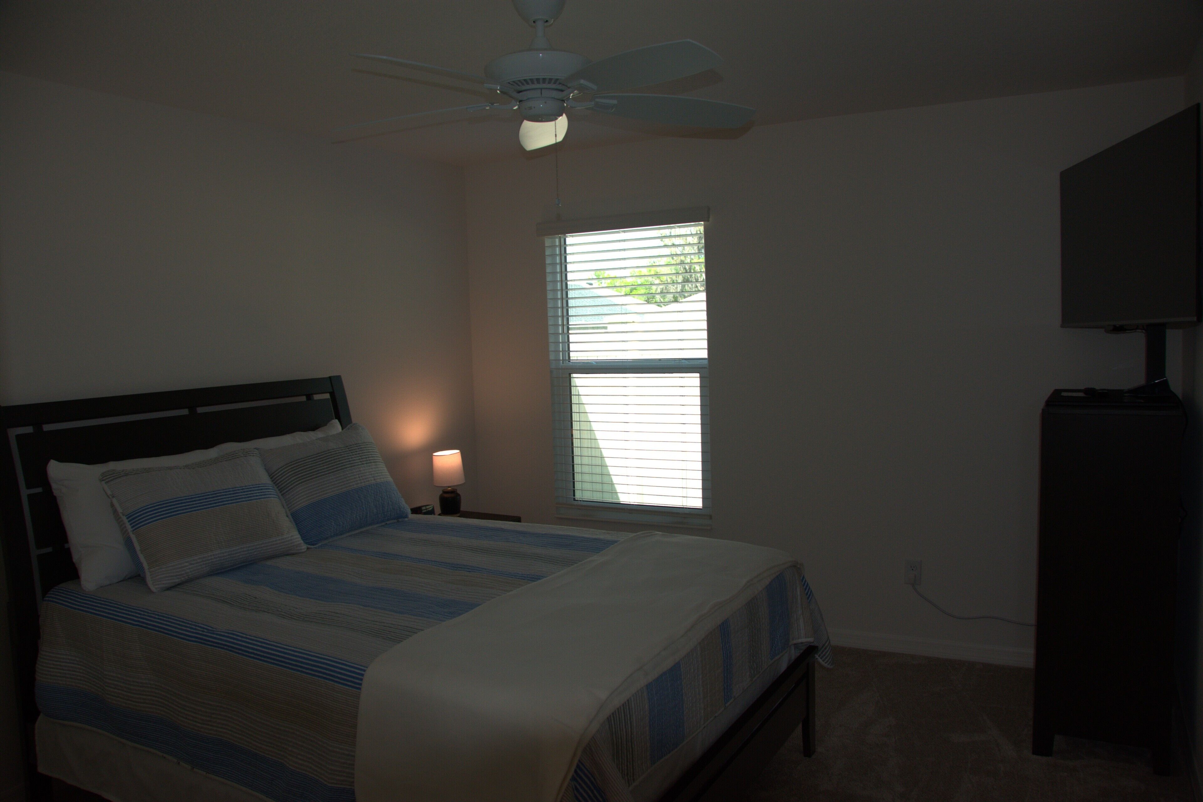 A bedroom with a bed, a ceiling fan, a window, and a TV on a stand.