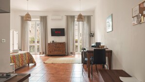 A spacious living area with a sofa, dining table, and chairs, a flat-screen TV mounted on the wall, and a large window with curtains. - Marina Square Open Space (Palermo)