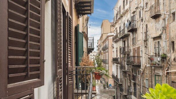 A narrow street with buildings on both sides, featuring balconies with plants and outdoor furniture. - Marina Square Open Space (Palermo)