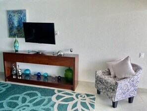 A living room with a flat-screen TV on a wooden console, decorative items, a patterned armchair, and a green patterned rug. - D-View & Relax Overlooking the Ocean (Puerto Vallarta)