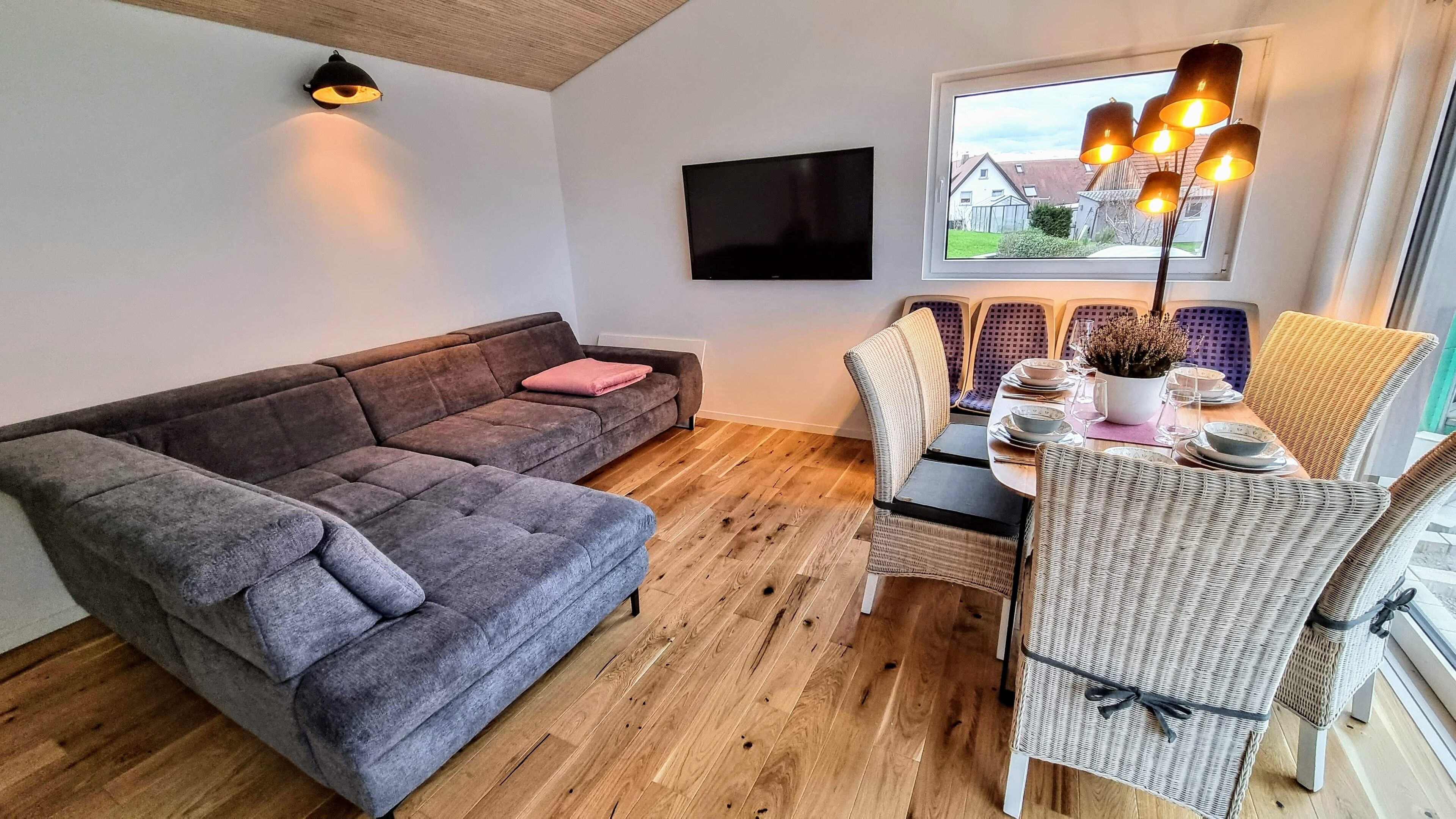 Ett vardagsrum med en grå soffa, ett matbord med stolar, en tv som är monterad på väggen och ett fönster med utsikt över hus.