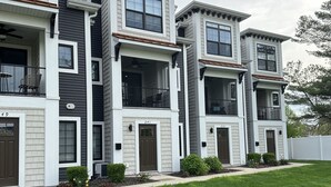 A row of modern multi-story townhouses with balconies, a well-maintained lawn, and a white picket fence. - Peaceful Condo (Grand Haven)