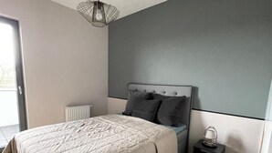 A modern bedroom with a large bed, a bedside table with a lamp, and a view of the outdoors through a window. - Poznan Surburb Apartament (Skórzewo)
