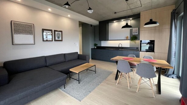 A modern living room with a dark grey sofa, a wooden coffee table, and a dining area with a wooden table and checkered chairs. - Poznan Surburb Apartament (Skórzewo)