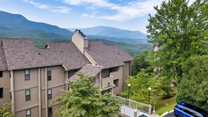 A multi-story building with a brick roof, surrounded by lush greenery and mountains in the background. - Deer Ridge Resort C207 - Amazing Mountain Views, Pool, and Tennis (Gatlinburg)