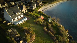 An aerial view of a coastal property with a large house, a well-maintained lawn, and a beach in the background.
