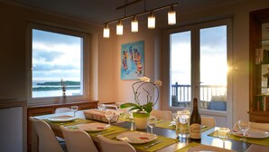 A dining area with a table set for four, large windows with a view, a painting on the wall, and a bottle of wine.