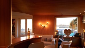 A cozy living area with a wooden ceiling, a bar with a wine bottle, and a view of the sea through large windows.