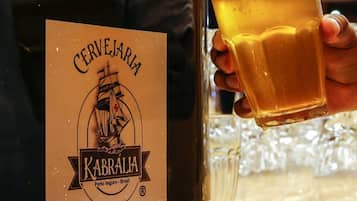 Eine Hand, die ein Glas Bier neben einer Flasche mit der Aufschrift 'Cerveja KABRALA Porto Seguro Brasil' hält.