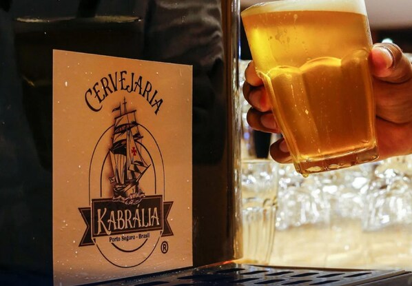 Käsi, jossa on lasi olutta, ja sen vieressä pullo, jonka etiketissä lukee: ”Cerveja KABRALA Porto Seguro Brasil”.
