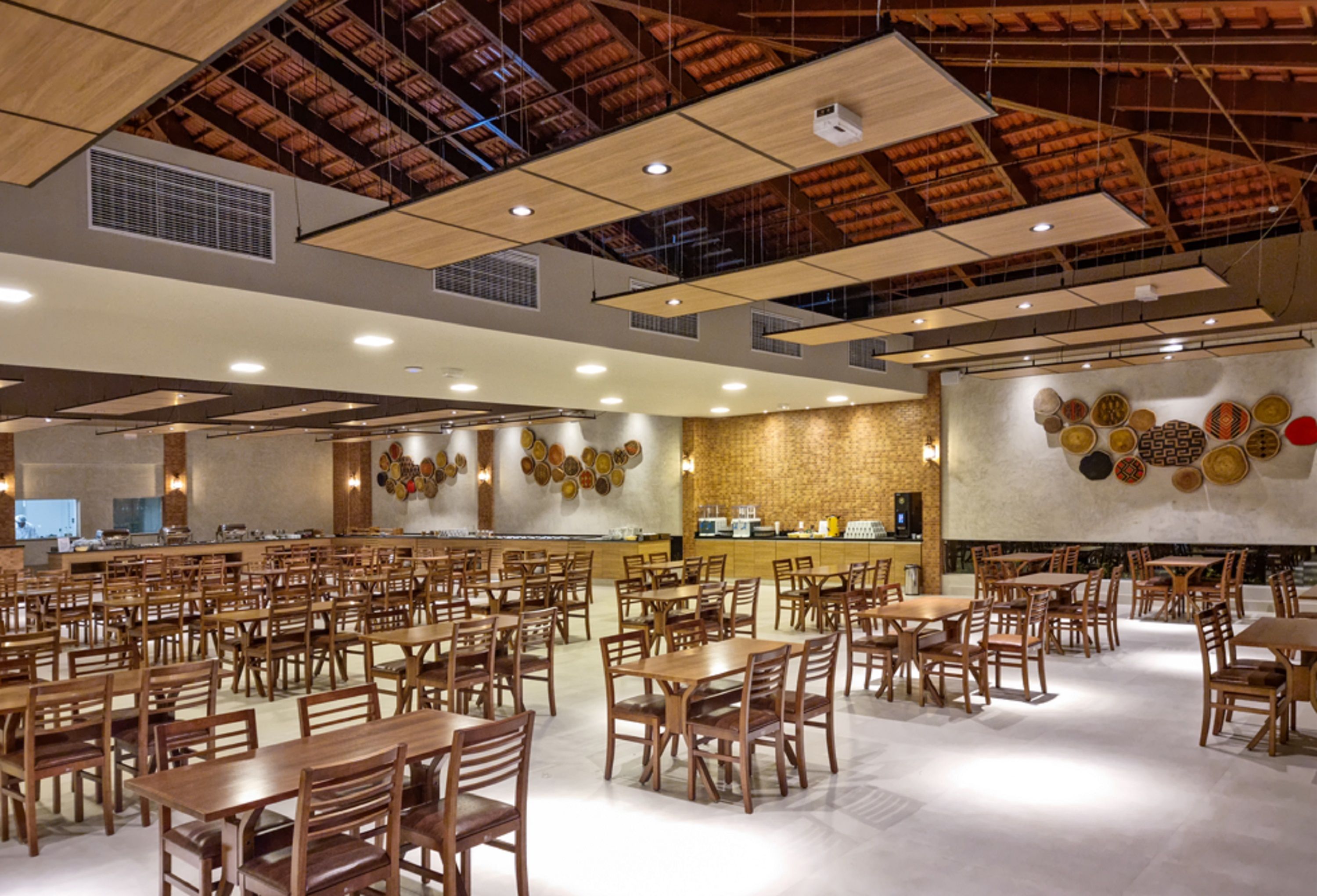 Uma ampla sala de jantar com várias mesas e cadeiras de madeira, teto alto com vigas aparentes e uma parede decorativa com padrões circulares.
