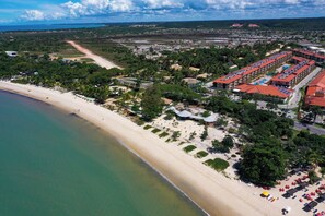 Vista aérea de un complejo turístico en la costa con playa de arena, zona de estacionamiento y un hotel con varias construcciones.