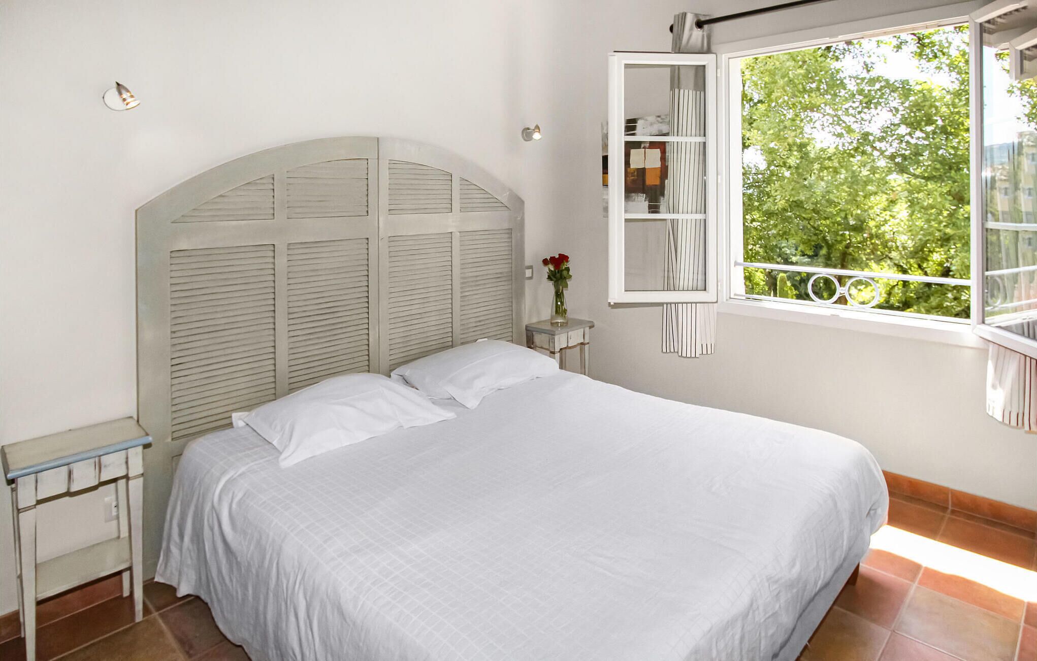 Ein Schlafzimmer mit einem Bett, einem Nachttisch, einem Fenster mit Blick auf Grünflächen und einem weißen Kopfteil.