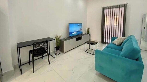 Una sala moderna con un sofá color turquesa, una mesa de comedor con sillas, un televisor sobre un mueble bajo y una ventana con persianas verticales.