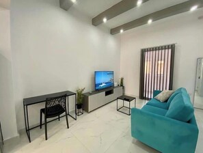 A modern living room with a teal sofa, a dining table with chairs, a TV on a stand, and a window with vertical blinds. - Seaside Apartment in Gzira (Il-Gżira)