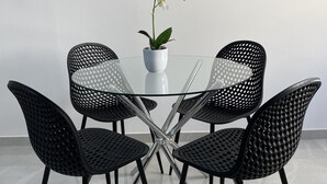 Uma mesa com tampo de vidro, quatro cadeiras pretas vazadas, uma orquídea branca em vaso e uma parede branca.