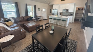 A spacious living area with a brown leather sofa, a dining table with chairs, and a kitchen with white cabinets and a bar area. - Fun New Lake House! (Canyon Lake)