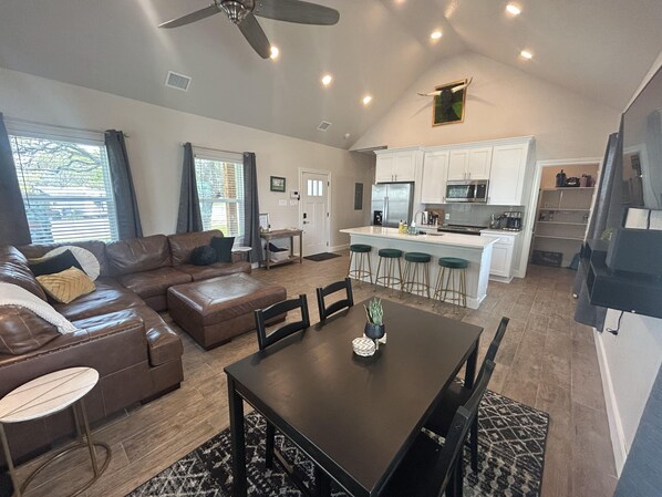 A spacious living area with a brown leather sofa, a dining table with chairs, and a kitchen with white cabinets and a bar area.