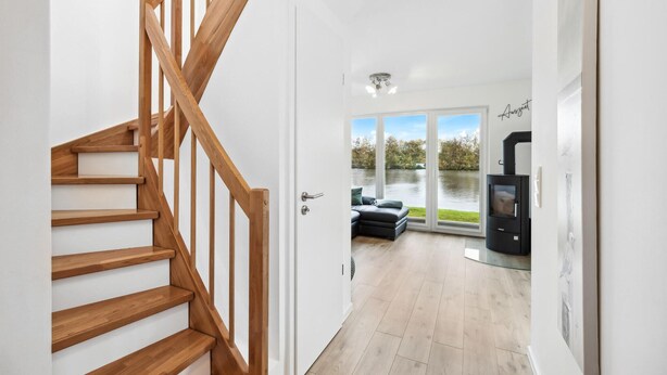 A modern interior with a wooden staircase, a cozy seating area with a view of a lake, and a wood-burning stove. - 2 bedroom gorgeous home in Tossens (Tossens)