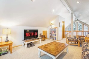 A spacious living room with a large flat-screen TV displaying 'NETFLIX', a bookshelf, a wooden coffee table, and a cozy seating area with a view of the outdoors.