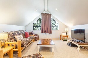 A cozy loft-style room with a sloped ceiling, a large window with curtains, a sofa, a coffee table, a TV, and a lamp. - Close to Ski + Snowboard | Games | Hiking Trails (Snoqualmie Pass)